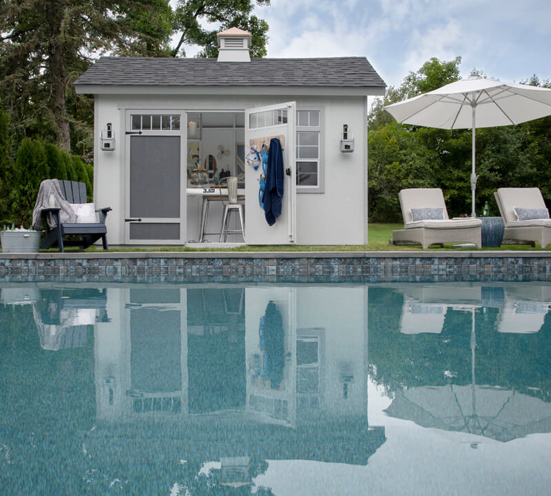White pool shed with gray doors next to in ground pool and big umbrella.