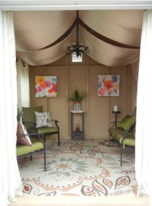 Social space She Shed: Four green chairs facing each other, each with a decorative throw pillow. Two art prints of flowers on one wall. Ceiling light fixture in center of room.